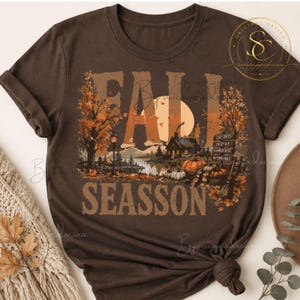 Op de afbeelding: Een donkerbruin t-shirt met korte mouwen en een grafische print van een herfstlandschap met een hut, pompoenen, bomen en een wegwijzer. De tekst luidt FALL SEASSON, met op de wegwijzer COZY DAYS, CRISP AIR, PUMPKIN SPICE en AUTUMN VIBES.