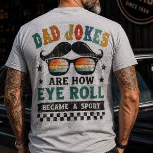 May include: A gray t-shirt with the text "DAD JOKES ARE HOW EYE ROLL BECAME A SPORT" in a retro font. The design includes a mustache, sunglasses, and stars. The shirt is short-sleeved.