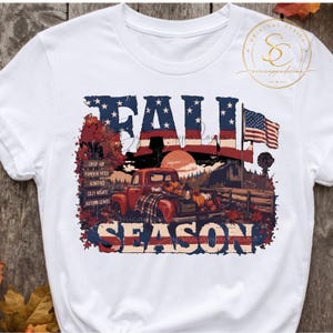 Op de afbeelding: Een wit t-shirt met een afbeelding met de tekst FALL SEASON. Het ontwerp bevat een rode pick-up truck vol pompoenen, een schuur, een windmolen en een wegwijzer met herfstthema's, alles tegen een achtergrond van de Amerikaanse vlag.