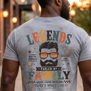 May include: A grey t-shirt with a graphic design. The design includes the word "LEGENDS" in orange, green, and blue. A bearded face wearing sunglasses is featured, along with the phrases "BUILT ON STRENGTH" and "DRIVEN BY LOVE". The words "FUELED BY FAMILY" are also present.