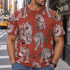 Op de afbeelding: Een t-shirt met korte mouwen in een diepe terracotta oranje kleur met een all-over print van luipaarden in verschillende houdingen. Het ontwerp bevat meerdere illustraties van gevlekte luipaarden, sommige zittend en sommige lopend, verspreid over de stof.