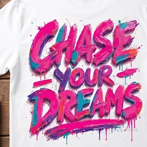 Op de afbeelding: Wit T-shirt met de tekst "CHASE YOUR DREAMS" in graffiti-stijl letters. De tekst is uitgevoerd in roze, paarse en blauwe tinten met druipende verfeffecten, wat zorgt voor een levendig en artistiek ontwerp.