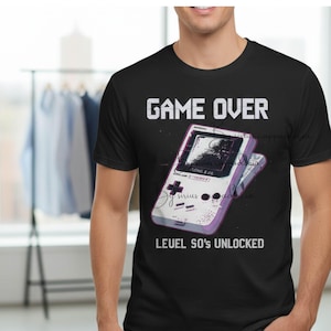 Op de afbeelding: Zwart t-shirt met een afbeelding van een retro handheld gameconsole. De tekst "GAME OVER" staat boven de console en "LEVEL 50'S UNLOCKED" eronder. De console is wit en paars.