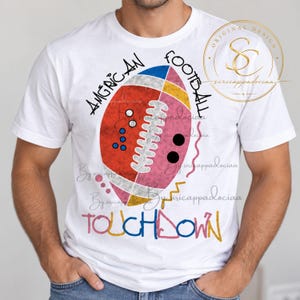Op de afbeelding: Een wit t-shirt met korte mouwen met een afbeelding van een American football verdeeld in rode, roze en gele secties. De tekst luidt "AMGRICAN FOOTBALL" en "TOUCHDOWN" in kleurrijke letters, vergezeld van kleine cirkels en zigzaglijnen.