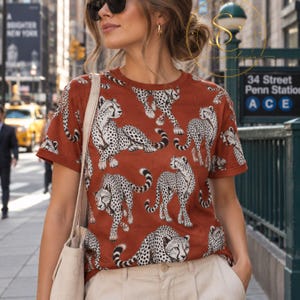 Op de afbeelding: Een roestkleurig T-shirt met korte mouwen en een all-over print van zwarte en witte cheeta's in verschillende houdingen, gecombineerd met een lichtbeige canvas draagtas en een lichtgekleurde broek.