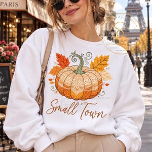 Op de afbeelding: Een wit sweatshirt met lange mouwen en een centrale afbeelding van een oranje pompoen met ingewikkelde geometrische patronen, omringd door oranje en gele herfstbladeren. De tekst "Small Town" is in bruin cursief schrift eronder gedrukt.