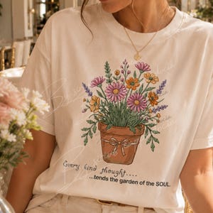 Op de afbeelding: Crèmekleurig T-shirt met een bloemenillustratie in een terracotta pot. Het ontwerp bevat roze, oranje en paarse bloemen met groene bladeren. De tekst luidt: "Every kind thought...tends the garden of the SOUL."
