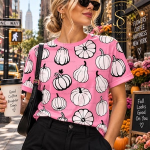 Op de afbeelding: Een roze T-shirt met korte mouwen en een herhalend patroon van witte pompoenen met zwarte omlijningen. Het shirt heeft een ronde hals en wordt gedragen met een zwarte broek en een zwarte schoudertas. Een witte koffiebeker wordt vastgehouden.