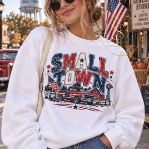 Könnte beinhalten: Ein weißes Langarm-Sweatshirt mit einem Grafikdruck, der den Text SMALL TOWN in fetten, abgenutzten Buchstaben zeigt. Das Design umfasst einen roten Pickup, eine amerikanische Flagge, eine rote Scheune und eine Windmühle in patriotischen Farben.