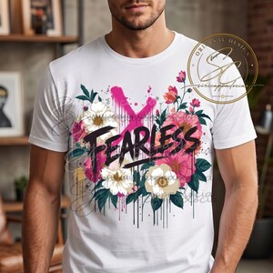 Op de afbeelding: Een wit T-shirt met korte mouwen en een centraal grafisch ontwerp van roze en witte bloemen met groene bladeren. Het woord FEARLESS is in dikke zwarte letters over het bloemstuk gedrukt, geaccentueerd door een felroze spuitverf X.