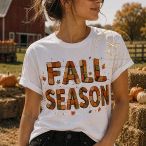 Op de afbeelding: Een wit T-shirt met korte mouwen en de tekst FALL SEASON in blokletters, opgebouwd uit een patchwork van oranje, bruine en groene vierkantjes. Het ontwerp is versierd met kleine verspreide herfstblaadjes en stippen in bijpassende seizoenskleuren.