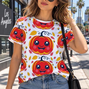 Op de afbeelding: Een wit t-shirt met korte mouwen voorzien van een all-over patroon van lachende oranje pompoenen met grote zwarte ogen en roze wangen, afgewisseld met kleine gele en rode herfstbladeren en eikels, gecombineerd met een blauwe spijkerbroek en een zwarte tas.
