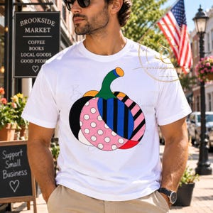 Op de afbeelding: Een wit t-shirt met korte mouwen en een grote, gestileerde pompoenafbeelding op de voorkant. De pompoen is verdeeld in secties met verschillende patronen, waaronder roze met witte stippen, blauw met zwarte verticale strepen en zwart.