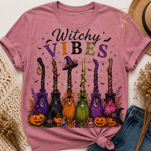 Op de afbeelding: Een mauve t-shirt met de tekst Witchy VIBES in kleurrijke letters boven een illustratie van zes versierde bezemstelen, een heksenhoed en drie pompoenen omringd door kleine sterretjes, vleermuizen en herfstbladeren.
