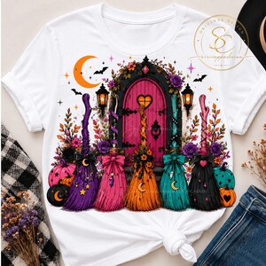 May include: A white t-shirt featuring a graphic print of five colorful broomsticks standing in front of a pink arched door. The design includes pumpkins, flowers, a crescent moon, bats, and lanterns. A circular logo with the text SC is at the top right.