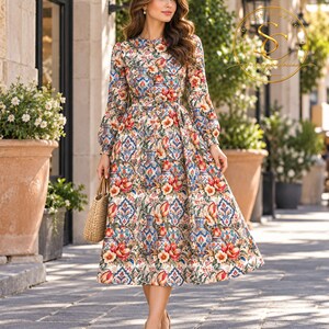 Op de afbeelding: Een midi-jurk met lange mouwen en een levendig patroon van blauw-witte vazen afgewisseld met rode en oranje tulpen. De jurk heeft een ronde halslijn, een getailleerde taille en een wijde rok. Een gevlochten strooien handtas wordt aan de zijkant gedragen.