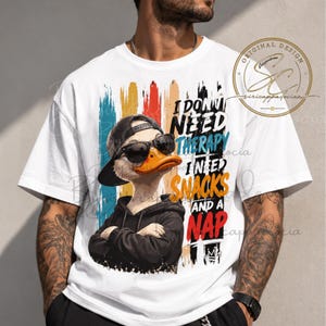Op de afbeelding: Een wit t-shirt met een afbeelding van een eend die een zwarte baseballpet, zwarte zonnebril en een zwarte hoodie draagt. De tekst luidt I DON'T NEED THERAPY I NEED SNACKS AND A NAP. Achtergrond met verticale penseelstreken in blauw, oranje en zwart.