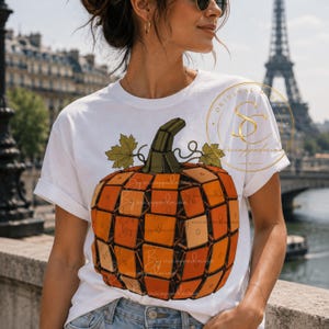 Op de afbeelding: Een wit t-shirt met korte mouwen en een grote afbeelding van een pompoen opgebouwd uit oranje en lichtoranje vierkante puzzelblokjes. De pompoen heeft een donkergroene steel en twee groene bladeren aan de bovenkant. Het ontwerp bevat een rond logo.