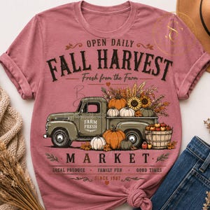 Op de afbeelding: Een mauve t-shirt met korte mouwen en een afbeelding van een vintage olijfgroene vrachtwagen met pompoenen en zonnebloemen. De tekst luidt: OPEN DAILY FALL HARVEST Fresh from the Farm FARM FRESH MARKET LOCAL PRODUCE FAMILY FUN GOOD TIMES SINCE 1987.