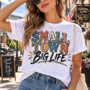Op de afbeelding: Een wit t-shirt met korte mouwen voorzien van de tekst "SMALL TOWN" in verweerde blokletters in blauw, groen en oranje, en de woorden "BIG LIFE" in een vet zwart handschrift, geaccentueerd door kleine kleurrijke horizontale lijnen.