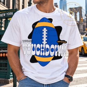 Op de afbeelding: Een wit t-shirt met korte mouwen en een grote blauwe en gele American football-afbeelding in het midden. Het woord TOUCHDOWN is gedrukt in grote witte letters met een zwarte omlijning. De tekst bevat RUNNING BACK, QUARTERBACK en LET'S PLAY.