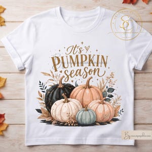 Op de afbeelding: Een wit t-shirt met korte mouwen en een grafische print van diverse pompoenen in zwart, oranje en lichtblauw, omringd door gedroogd loof. De tekst "It's PUMPKIN season" staat in een bruin script en schreeflettertype boven de pompoenen.