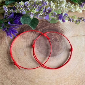 May include: Two red string bracelets tied together in a circle. The bracelets are made of thin red string and have knots at the ends.