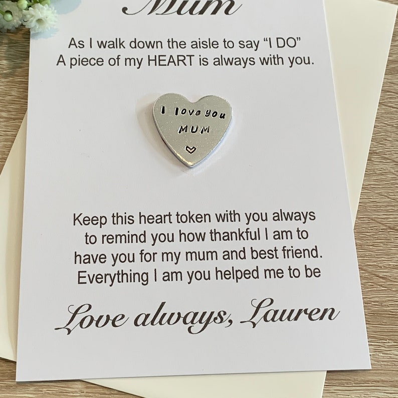 May include: A white card with a silver heart charm attached. The card reads "As I walk down the aisle to say 'I DO' A piece of my HEART is always with you." The heart charm reads "I love you MUM" and has a small heart at the bottom. The card is signed "Love always, Lauren".