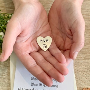 May include: A silver heart-shaped charm with the word "MUM" and a small angel design. The charm is being held in the palm of a hand.
