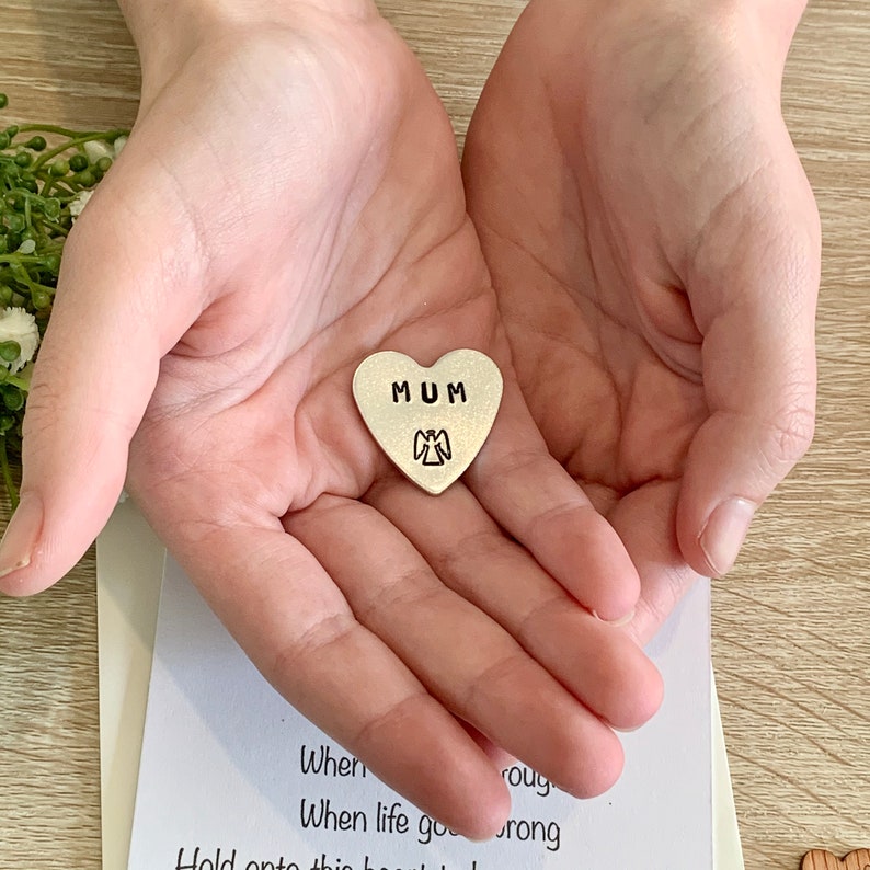 May include: A silver heart-shaped charm with the word "MUM" and a small angel design. The charm is being held in the palm of a hand.