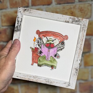 May include: Watercolor painting in a distressed white frame. The artwork depicts a whimsical creature with moth wings reading a book in bed. A candle and a cup sit on a nightstand. The bed and nightstand are painted in shades of red and brown.
