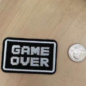 May include: Black and white embroidered patch with the text "GAME OVER" in a white pixelated font.