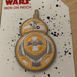 May include: A white and gold Star Wars iron-on patch featuring a droid character. The patch is on a white background with black splatter paint.