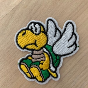 May include: An embroidered patch featuring a yellow and green cartoon turtle with white wings. The turtle is sitting with its wings spread out and has a closed eye.