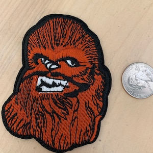 May include: Embroidered patch of Chewbacca from Star Wars. The patch is orange and brown with a black outline. It features Chewbacca's face with a white smile and black eyes.