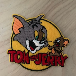 May include: Tom and Jerry cartoon character embroidered patch with a yellow background and red lettering. The patch features Tom the cat looking at Jerry the mouse.