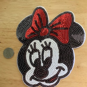 May include: A sequined Minnie Mouse patch with a red bow, black ears, and a white face. The patch is made of small, shiny sequins and is designed to be sewn or ironed onto fabric. A quarter is next to the patch for size comparison.