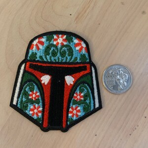 May include: Embroidered patch featuring a stylized helmet design with floral patterns. The helmet has a black visor, red and green accents, and white and orange flowers on a light blue background. A quarter coin is next to the patch.