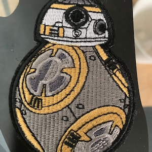 May include: A gray and yellow embroidered patch of a Star Wars BB-8 droid. The droid is round with a white dome on top. The dome has a black eye and a white and black stripe.