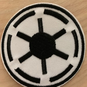 May include: A white embroidered patch with a black Imperial symbol from Star Wars. The symbol is a circle with a five-pointed star inside.