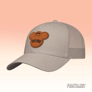 May include: A light beige trucker hat featuring a tan leather-like patch on the front. The patch is shaped like a mouse head with a small crown on top and includes the text YOUR NAME printed in black capital letters across the center.