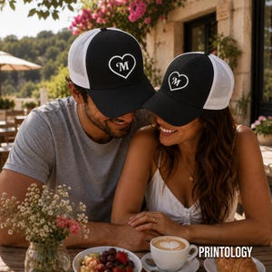 May include: Two black and white trucker hats featuring a white heart graphic with the letter M centered inside. The hats have a solid black front panel and a white mesh back panel. The design is identical on both hats, showcasing a structured crown.