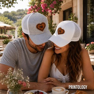 May include: A heather gray trucker hat and a white trucker hat, both featuring a brown leather-like heart patch on the front panel with the letter M embossed in the center. Each hat has a mesh back panel and a curved brim for a classic look.