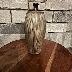 May include: A brown and beige ceramic vase with a dark brown lid. The vase has vertical textured lines and sits on a round wooden surface. The background features a stone wall.