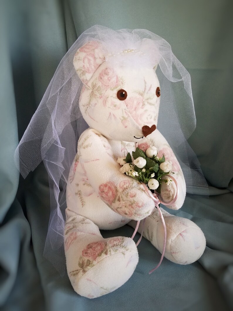 K&ouml;nnte beinhalten: Ein wei&szlig;er Teddyb&auml;r, der einen wei&szlig;en Schleier tr&auml;gt und einen Strau&szlig; wei&szlig;er Blumen h&auml;lt. Der B&auml;r ist in einem Stoff mit floralem Muster mit rosa Rosen gekleidet.