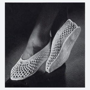 May include: A black and white photograph of a pair of crocheted slippers. The slippers are made with a simple stitch and have a pointed toe. The photo is taken from a low angle, showing the slippers on a person's feet.