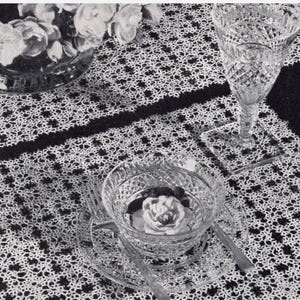 May include: A white crocheted table runner with a delicate floral pattern. The runner is laid out on a black surface with a glass bowl and a glass goblet.