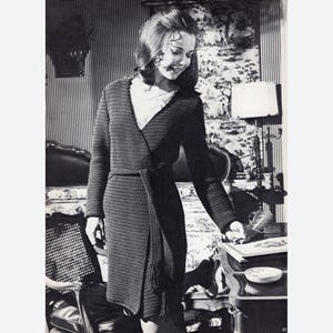 May include: A black crochet wrap with a tie belt. The wrap is worn by a woman standing in a room with a bed and a side table.
