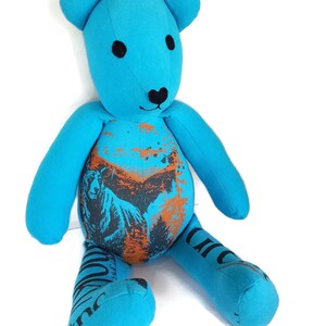 K&ouml;nnte beinhalten: Ein blauer Teddyb&auml;r mit schwarzen Augen und einer schwarzen Nase. Der B&auml;r sitzt auf einem wei&szlig;en Hintergrund. Das Fell des B&auml;ren ist aus recyceltem Stoff und hat ein bedrucktes Design mit einem B&auml;ren und B&auml;umen in Orange und Schwarz.