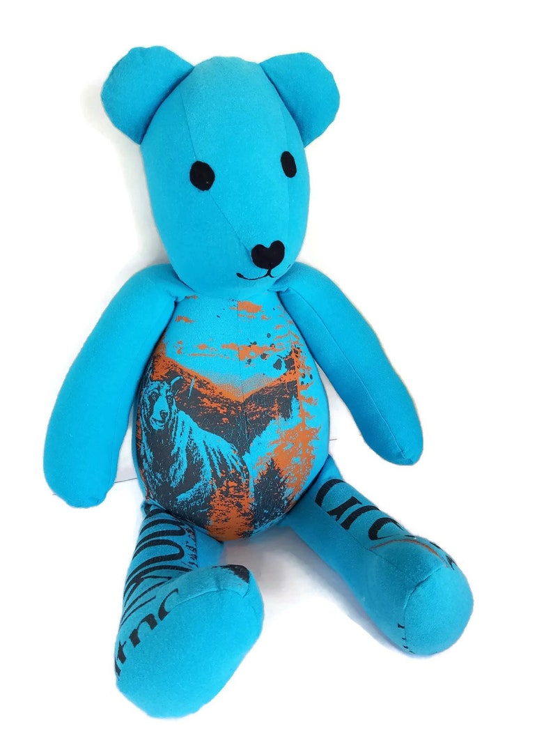 K&ouml;nnte beinhalten: Ein blauer Teddyb&auml;r mit schwarzen Augen und einer schwarzen Nase. Der B&auml;r sitzt auf einem wei&szlig;en Hintergrund. Das Fell des B&auml;ren ist aus recyceltem Stoff und hat ein bedrucktes Design mit einem B&auml;ren und B&auml;umen in Orange und Schwarz.